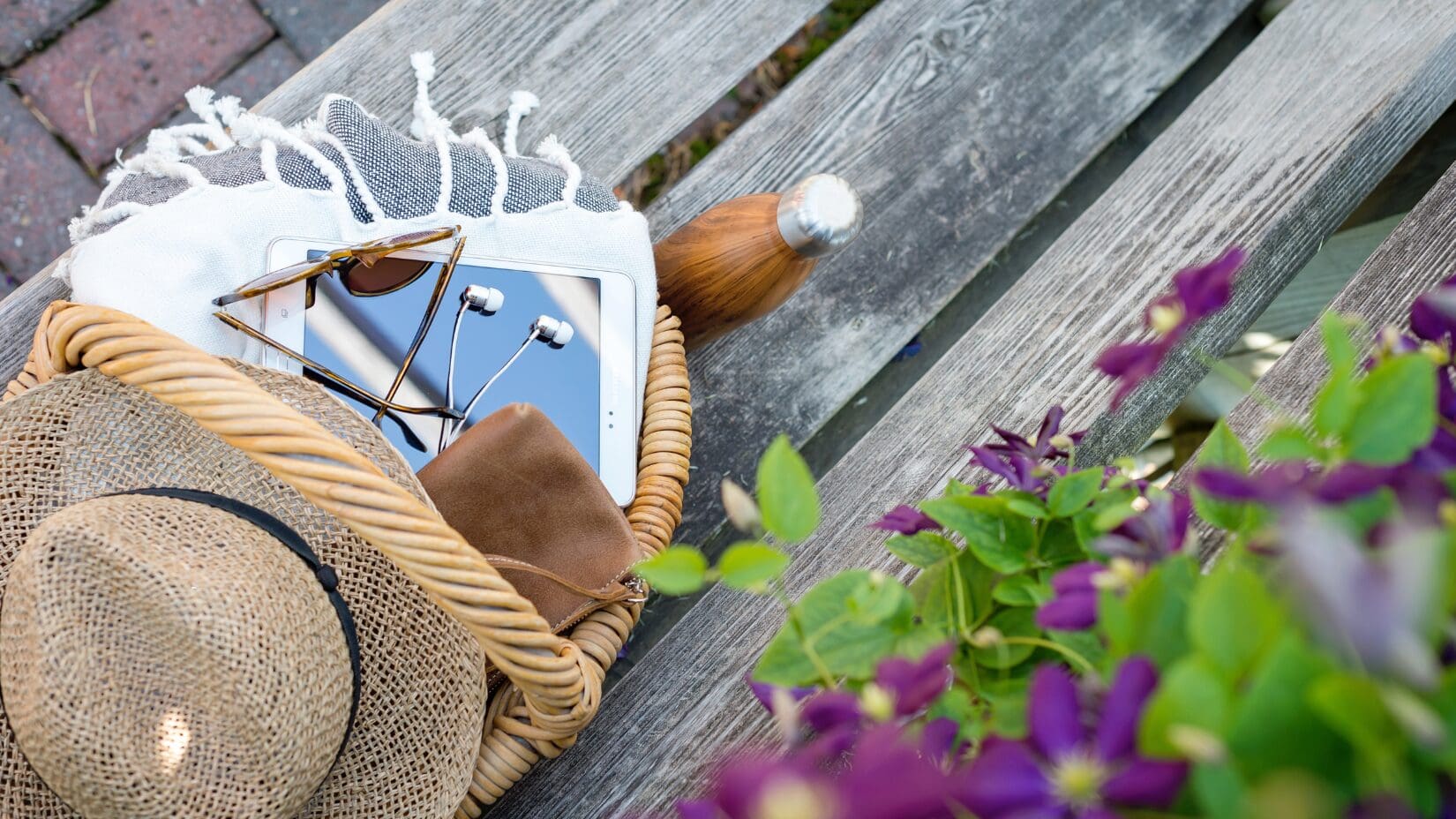 Shows an ipad, straw hat and earpods sitting on a bench with some purple flowers behind it