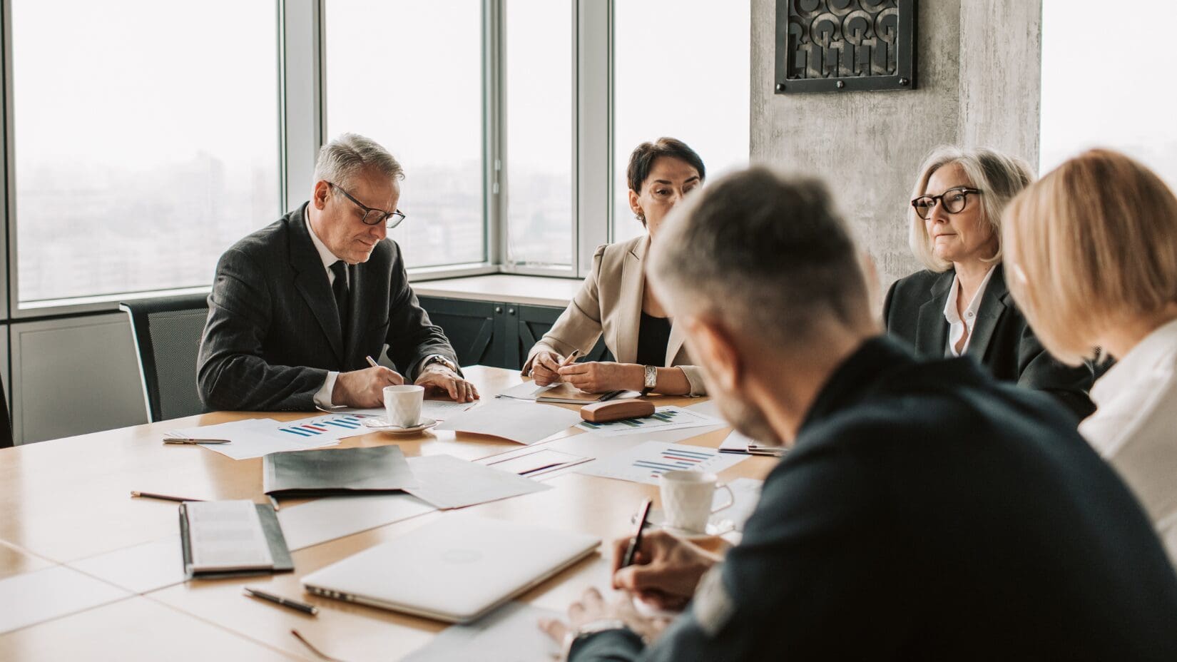 Business men and women sitting at a table in a conference room. Some are writing, there are documents on the table.