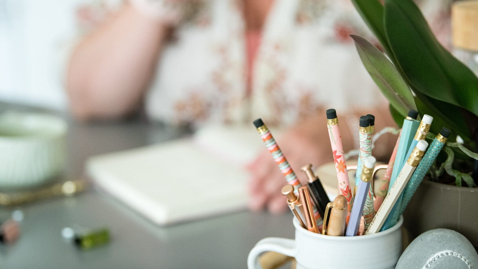 blurred photo shows woman writing in a notebook. there is a pencil cup and houseplant in the right hand side of the photo